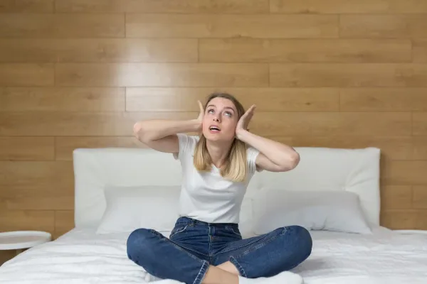 Žena sedí na posteli, zakrýva si uši a trápi ju kročajový hluk zo susedného bytu.